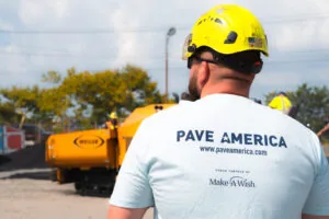 paving contractor in safety helmet facing away from camera, back of his shirt says "pave america, proud partner of make a wish"