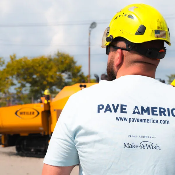 paving contractor in safety helmet facing away from camera, back of his shirt says "pave america, proud partner of make a wish"